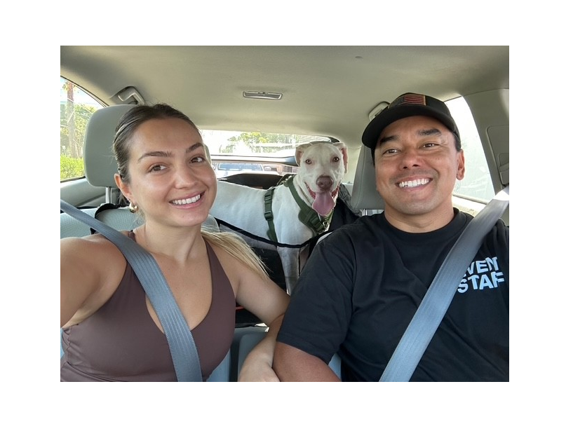 A couple smiles in a car, with their white dog sticking its head between them, all wearing seatbelts.