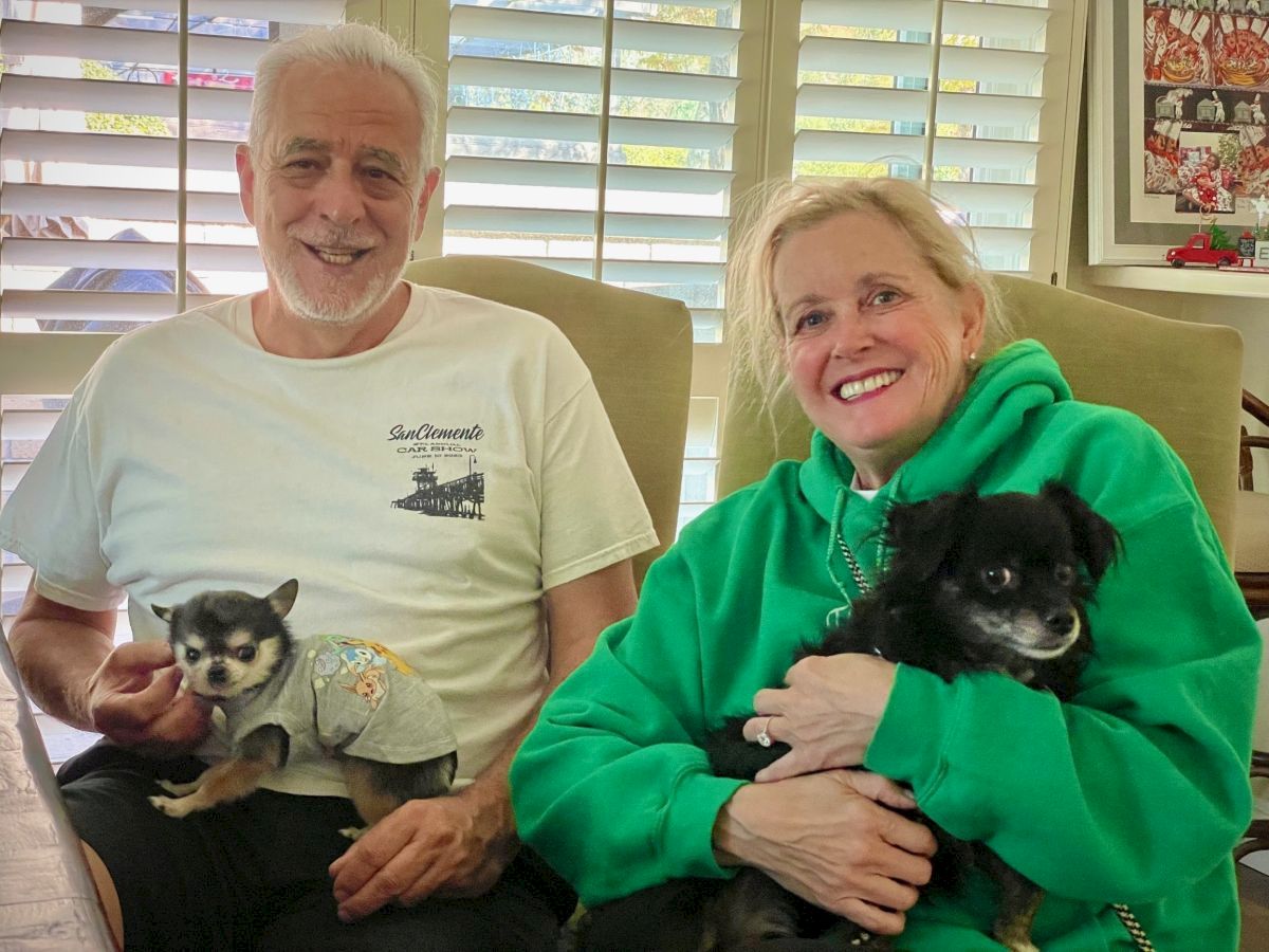 Two seniors sit on a couch with small dogs; man in white tee, woman in bright green hoodie, both smiling warmly at the camera.