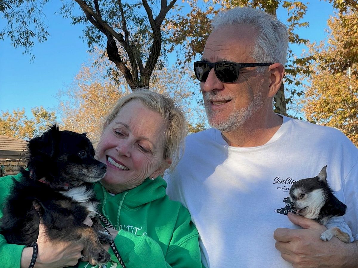 Two smiling adults hold small dogs outdoors on a sunny day, wearing casual clothes; a tree-lined park background.
