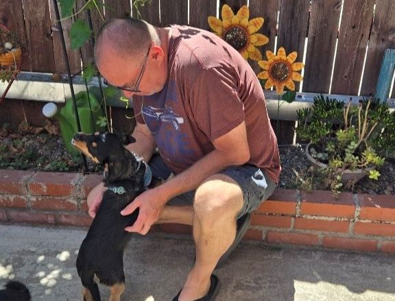 A man kneels in a backyard, gently petting a black dog while sunflowers and plants decorate the brick wall behind them.