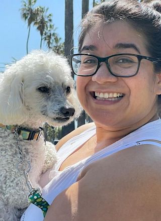 A smiling woman with glasses cuddles a fluffy white dog outdoors with palm trees behind them.