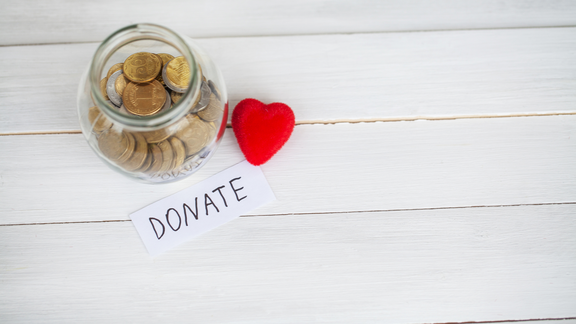 A glass jar filled with coins sits next to a red heart and a note that reads &ldquo;DONATE&rdquo; on a white wooden surface.