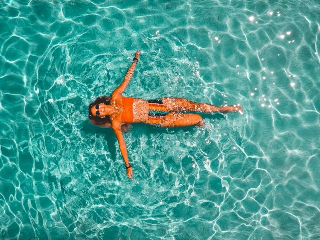 A person in a bright bikini floats face up on crystal-clear turquoise water, arms and legs relaxed as sunlight dances on the rippling surface.