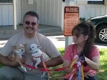 A man and a woman sit on the grass with two small white dogs, each dog wearing a red leash; they&rsquo;re outdoors near a building and a car, smiling.