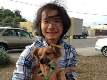 A smiling person holding a small brown dog with a green bandana outdoors by the road, in a casual plaid shirt.