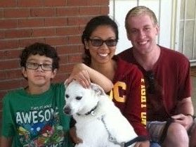 Three people and a white dog posing on a porch, smiling at the camera, with a brick wall in the background.